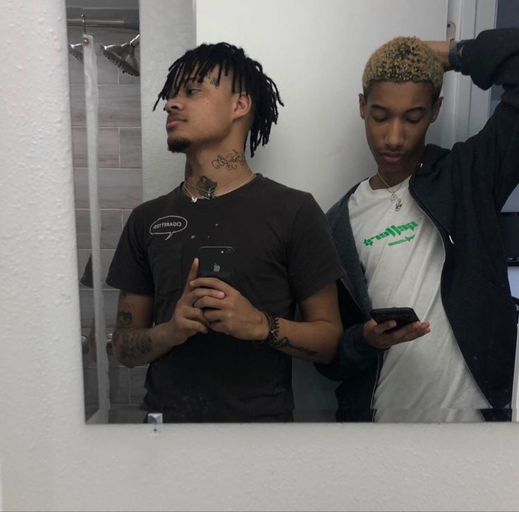 two young men standing in front of a mirror looking at their cell phones and taking a selfie