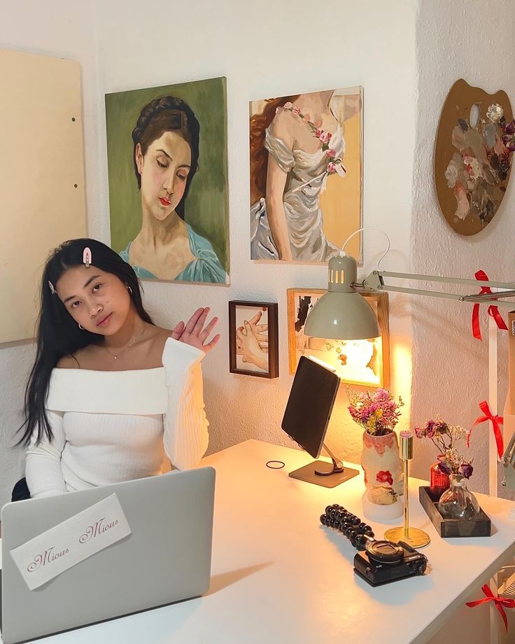 a woman sitting at a desk in front of a laptop computer with paintings on the wall behind her