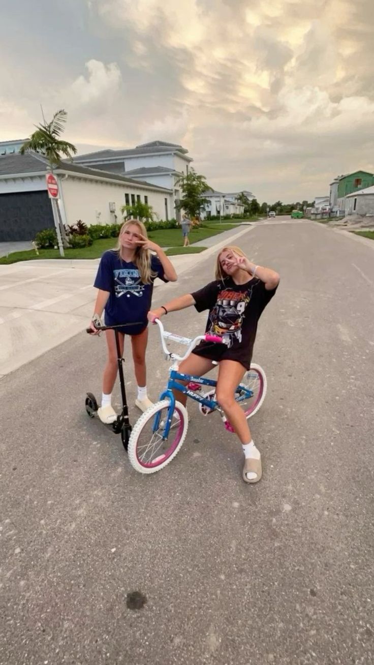 two girls are posing on their bikes in the middle of an empty street with houses behind them