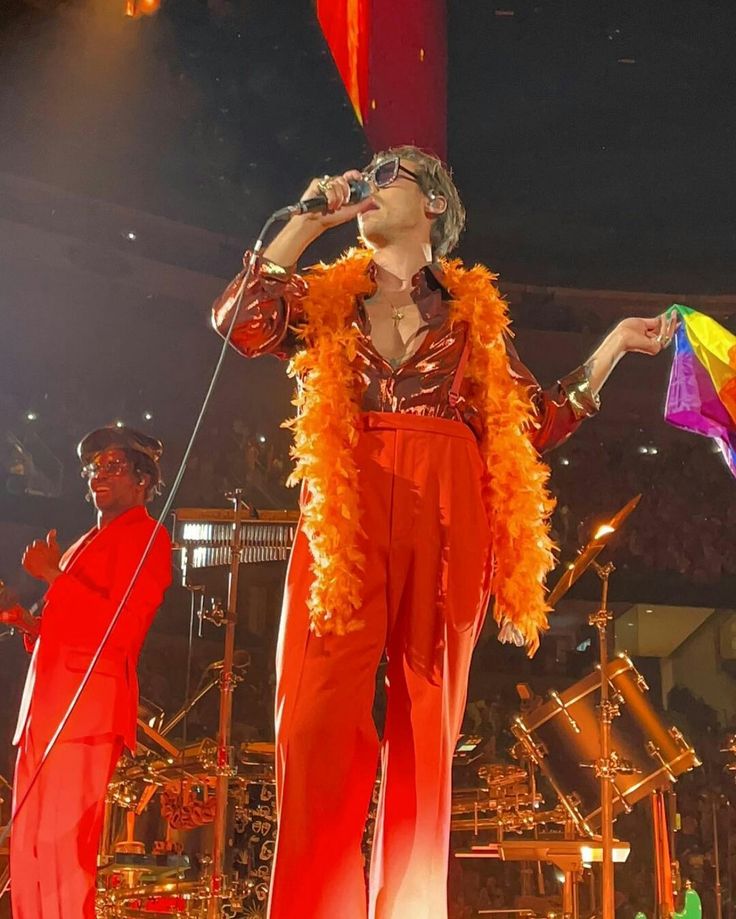 a man in an orange outfit on stage with his hands up to his head and microphone