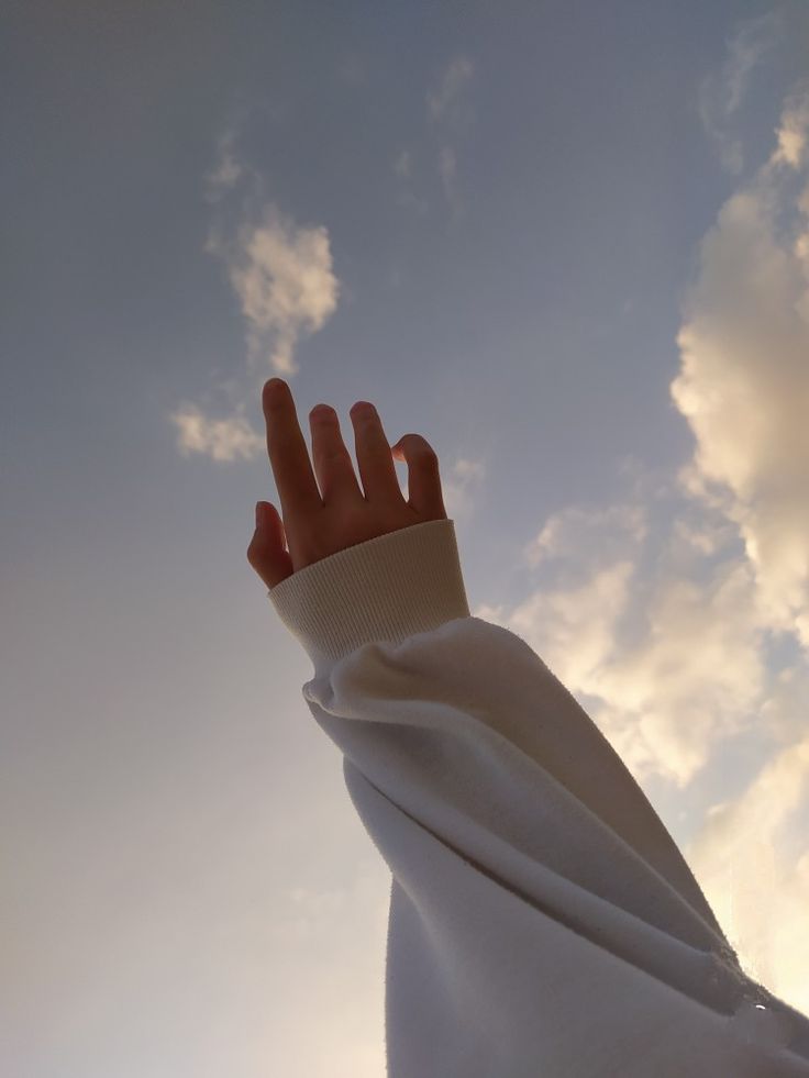 a person's hand reaching up into the air with clouds in the sky behind them