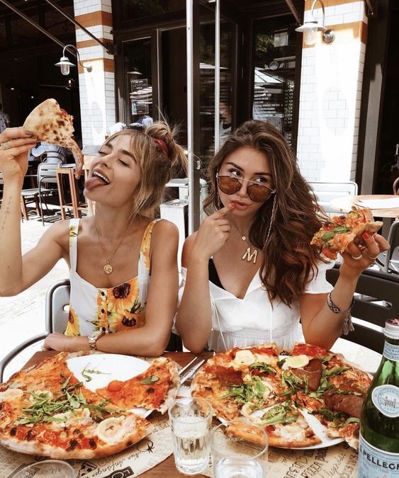 two beautiful women sitting at a table with pizza and drinks in front of them,