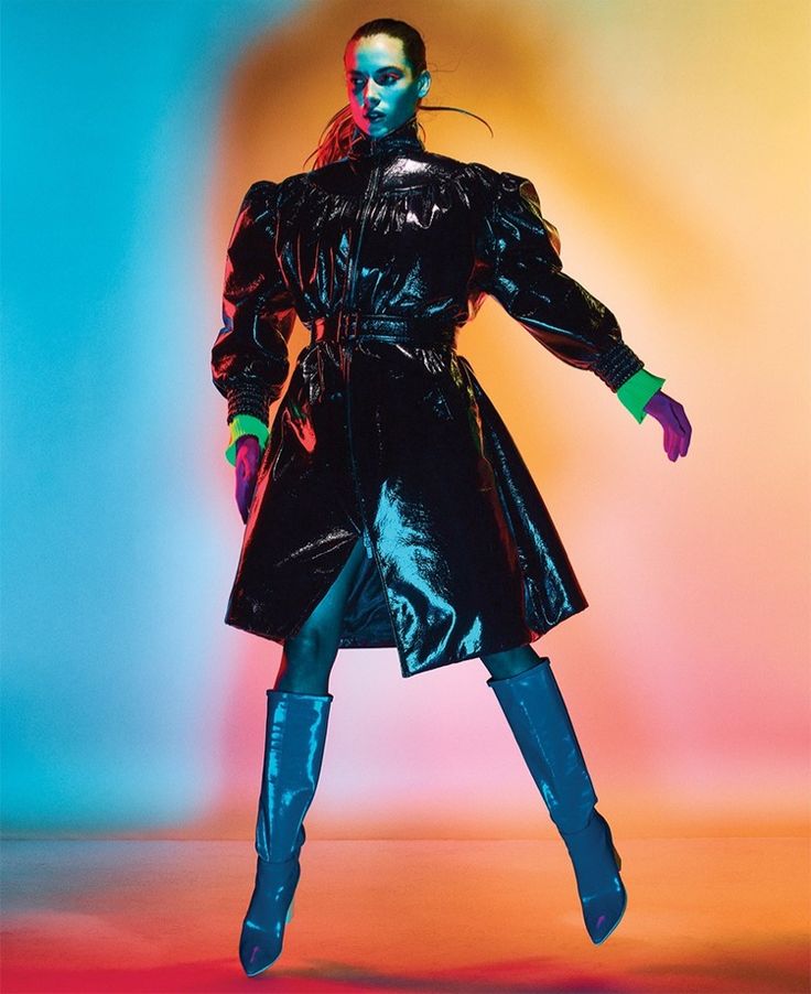 a woman in black leather coat and knee high boots standing against a colorful background with her arms outstretched