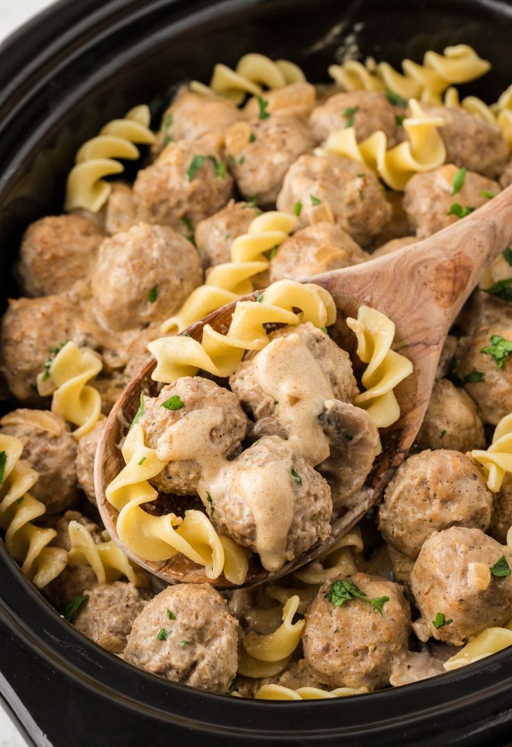 a wooden spoon in a slow cooker filled with pasta and meatballs, ready to be eaten