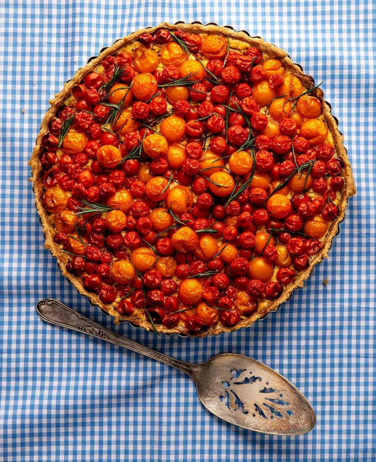 a blue and white checkered table cloth with a pie topped with cherries next to a spoon