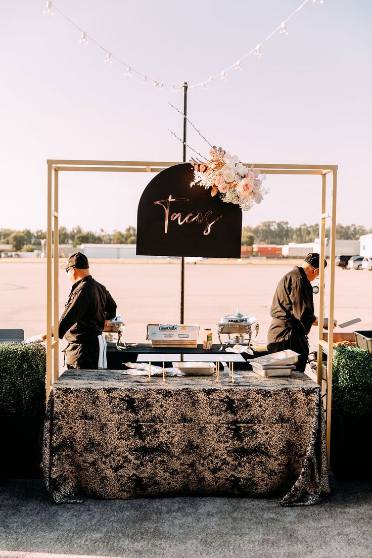 two men standing at a table with food in front of it and a sign that says tacos