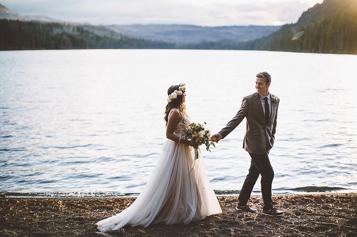 a bride and groom holding hands by the water at their lakeside wedding ceremony in british columbia