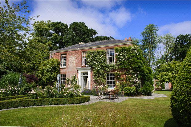 an old brick house surrounded by lush green trees and shrubs with a bench in the front yard