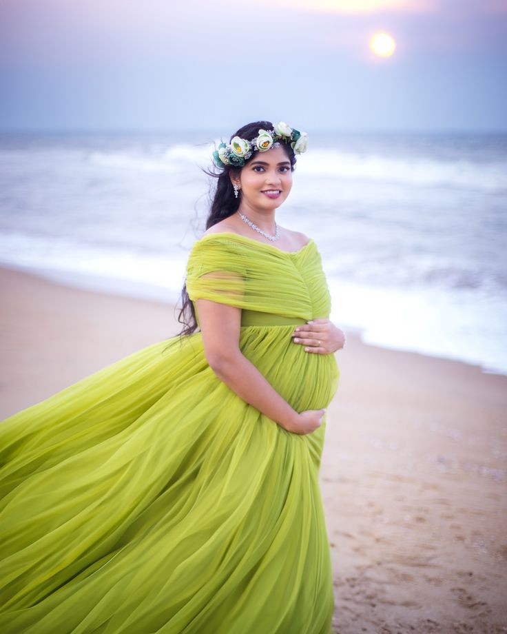 a pregnant woman standing on the beach in a green dress with flowers in her hair