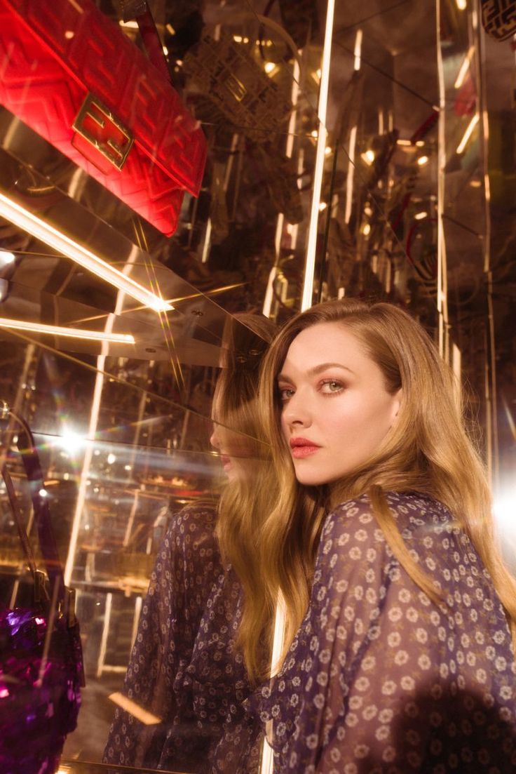 a woman standing in front of a mirror with lights behind her and bags hanging from the ceiling