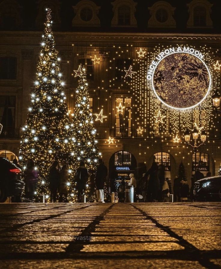 christmas trees are lit up in front of a building with lights on the sides and around them