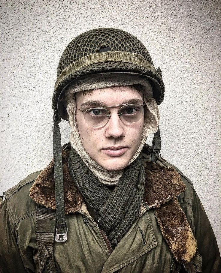 a young man wearing an army uniform with a helmet on his head and glasses in front of a white wall