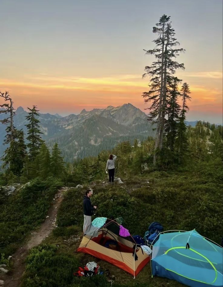 two people standing on the top of a mountain with tents set up in front of them