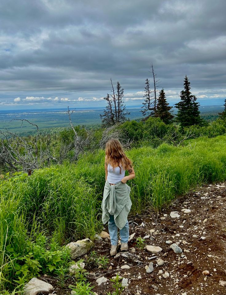 a woman standing on top of a lush green hillside next to a forest filled with lots of trees