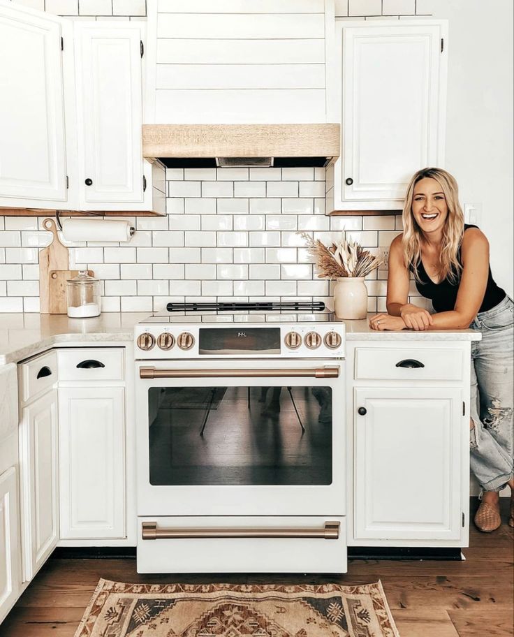 a woman leaning on the counter in a kitchen with white cabinets and an area rug