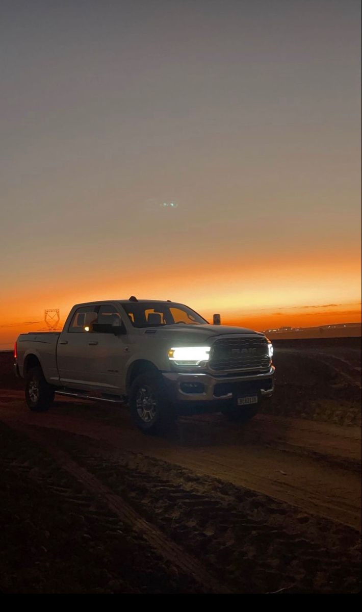 a white truck driving down a dirt road at sunset with the sun setting in the background