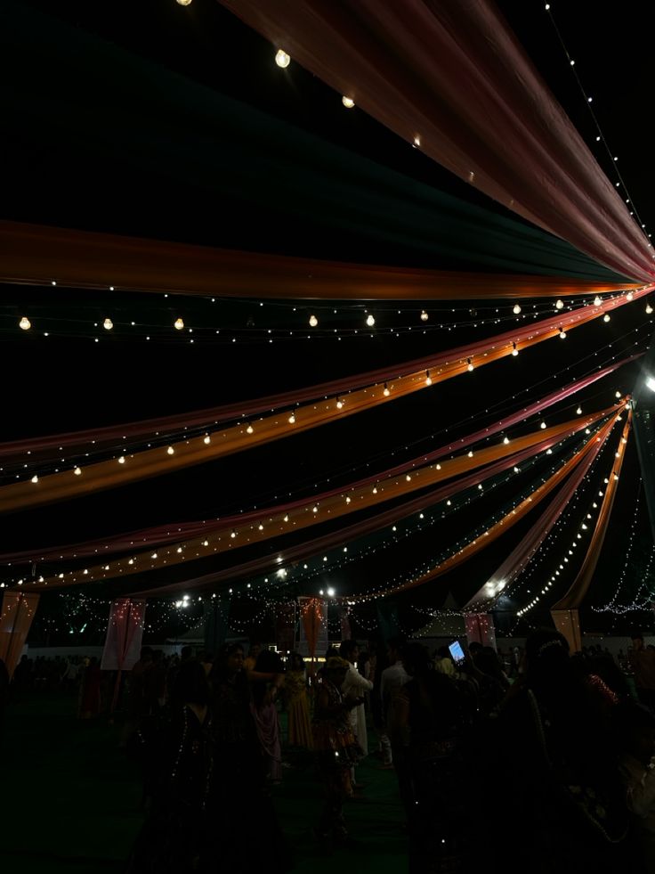 people are standing under the lights at night in front of a large tent with many strings hanging from it