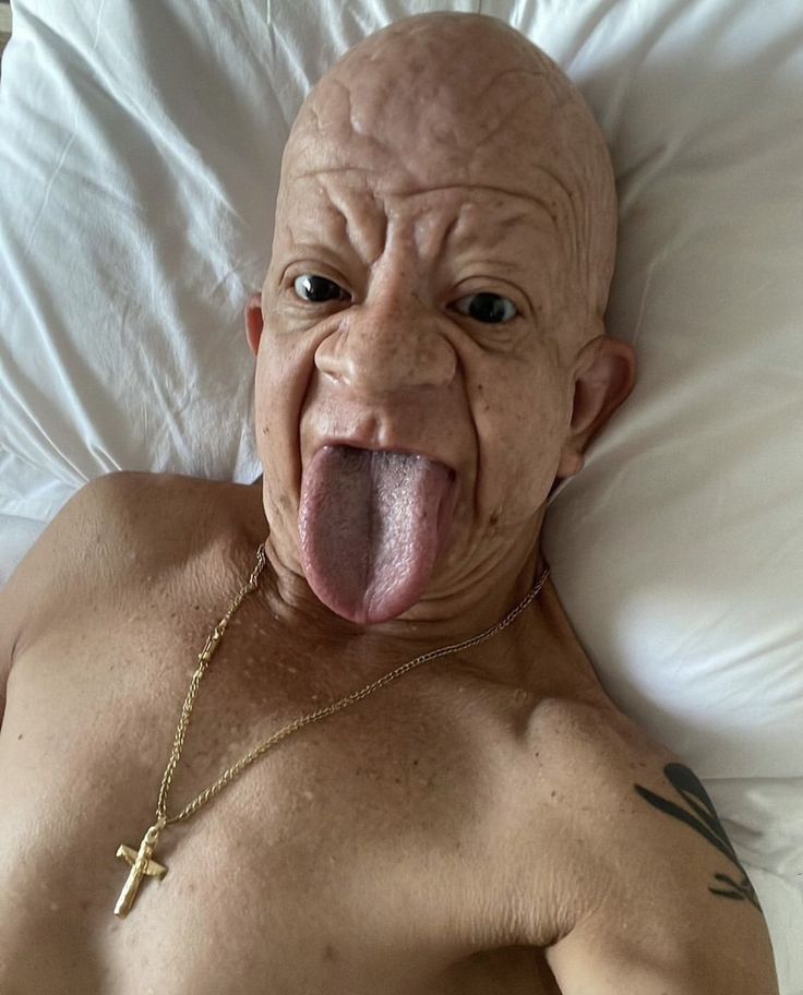 an old man sticking his tongue out while laying in bed with a cross on it's chest