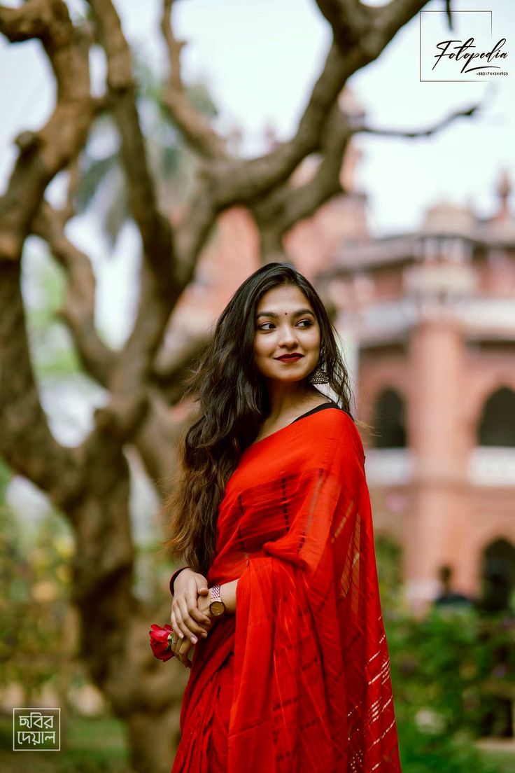 a woman in a red sari posing for a photo with trees and buildings behind her