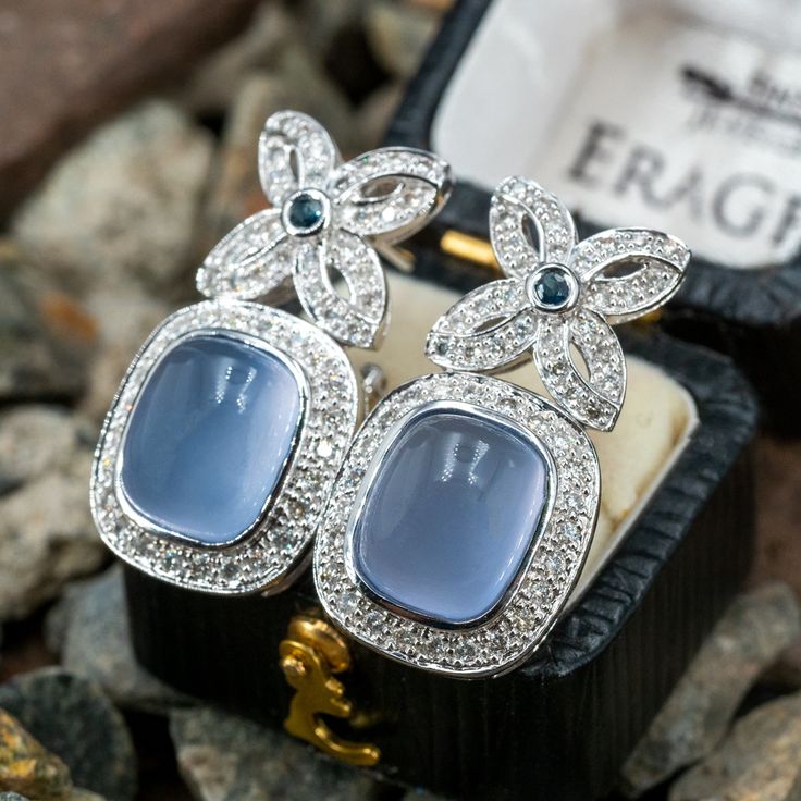 These classy dangle style pierced earrings are each accented with one (1), bezel set, cushion cabochon cut natural blue chalcedony, fifty-two (52), bead set, round brilliant cut diamonds and one (1), bezel set, round mixed cut natural blue sapphires. The earrings measure 28.0mm X 14.8mm and are finished with omega style backs. Elegant Chalcedony Drop Earrings, Formal Chalcedony Cabochon Jewelry, Formal Cabochon Chalcedony Jewelry, Elegant Chalcedony Jewelry For Anniversary, Elegant Blue Cushion Cut Earrings, Elegant Chalcedony Jewelry With Gemstone Accents, Elegant Blue Cabochon Earrings, Bead Set, Green Quartz