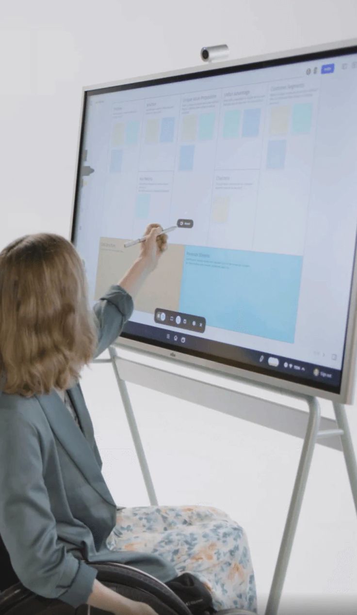 a woman is sitting in front of a large whiteboard and writing on the board