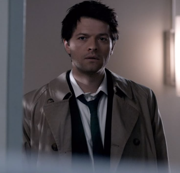 a man in a trench coat and tie looking at the camera while standing in front of a mirror