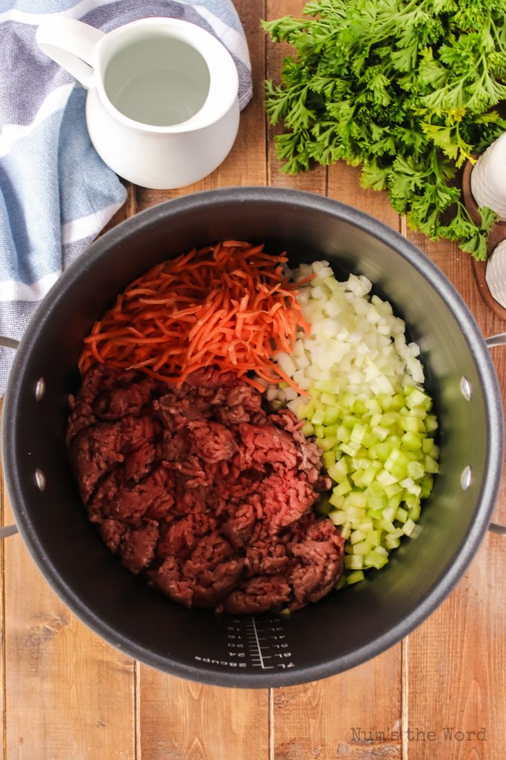 meat, onions and carrots in a pot on a wooden table next to some parsley