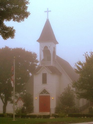 a white church with a red door and cross on the steeple in foggy weather