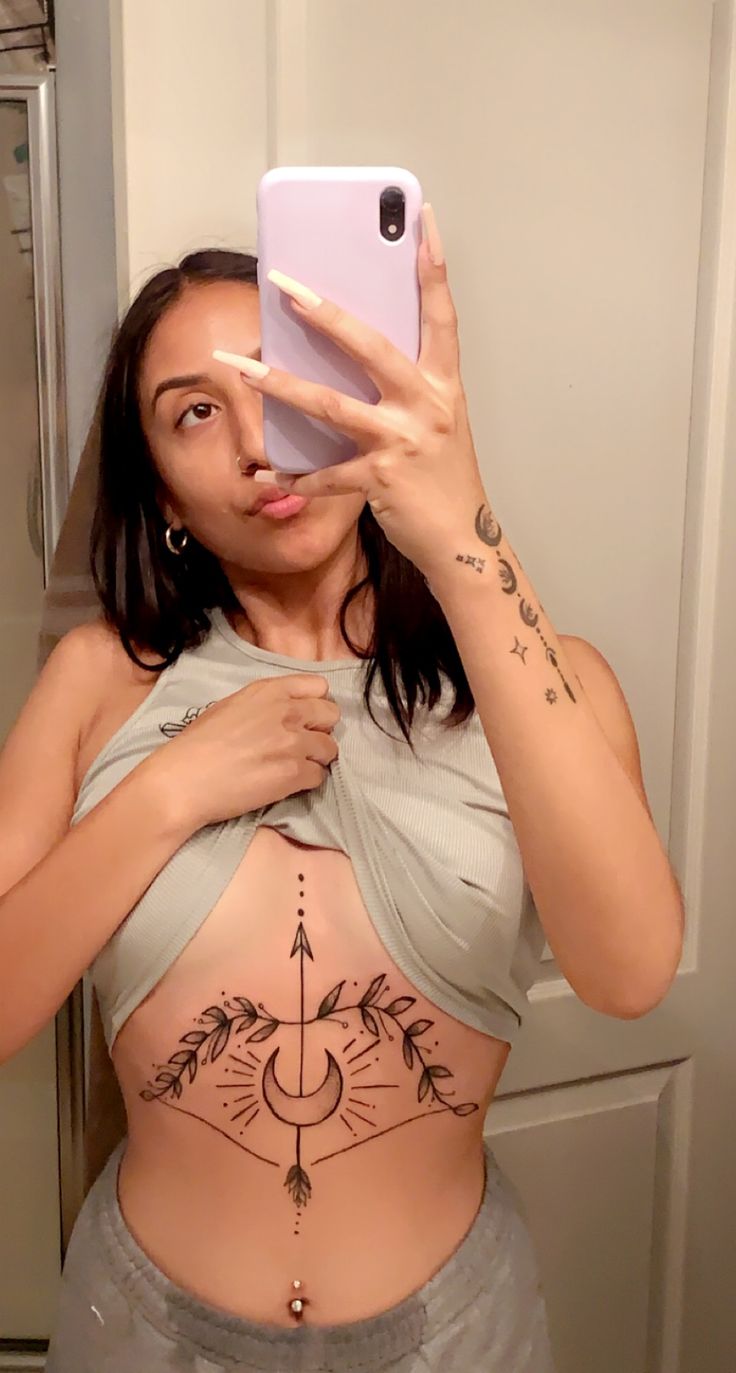 a woman taking a selfie with her cell phone in front of her stomach tattoo