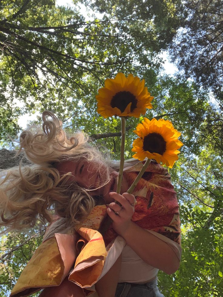 a woman with blonde hair holding a sunflower in her hand and looking up at the sky