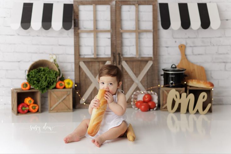 a baby is sitting on the floor eating a large baguette in front of some vegetables