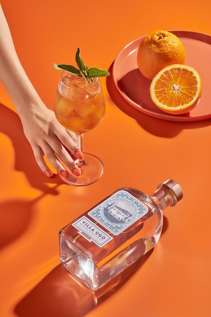 a person holding a glass with an orange in it next to a bottle of alcohol