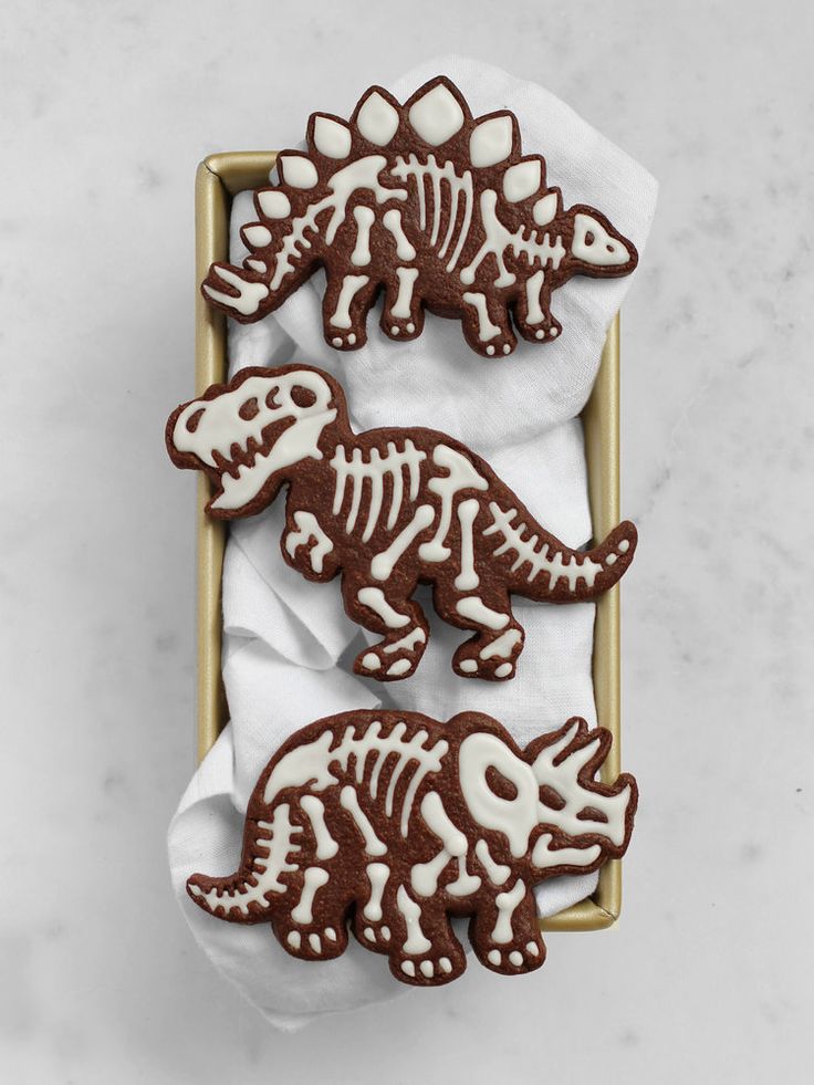 three decorated cookies in the shape of dinosaurs on a tray with white napkins and chocolate icing