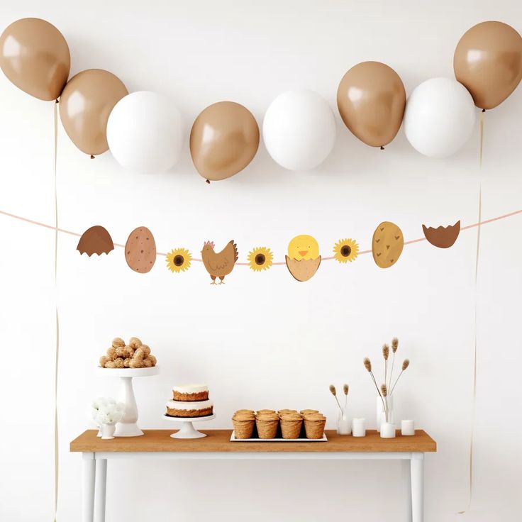 a table topped with cakes and balloons next to a wall filled with white, brown and gold balloons