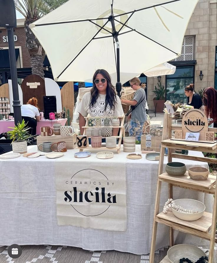 a woman standing behind a table with cups on it and an umbrella in the background