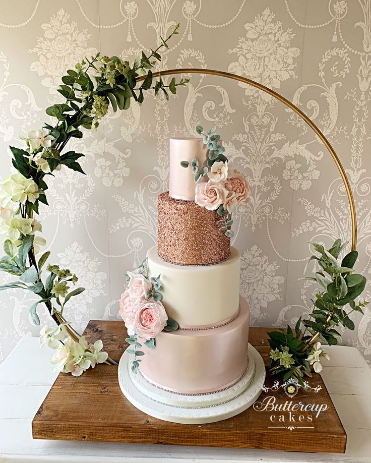 a three tiered cake with pink flowers and greenery on top, sitting on a wooden table