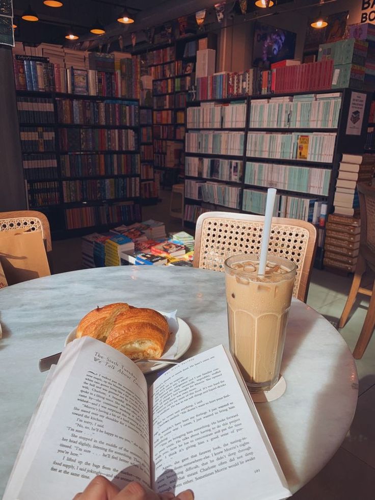 a person is reading a book at a table with a cup of coffee and croissant