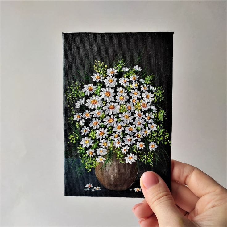 a painting of daisies in a vase is being held by a person's hand