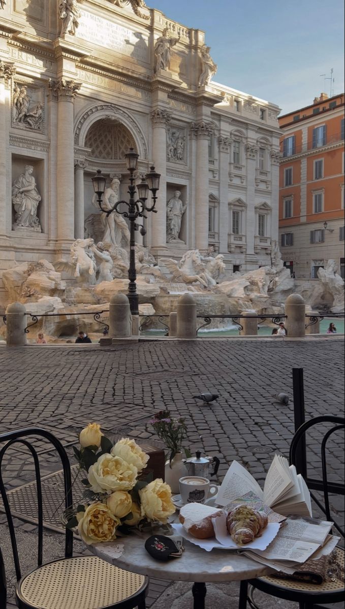 an outdoor table with food on it in front of a fountain and building behind it