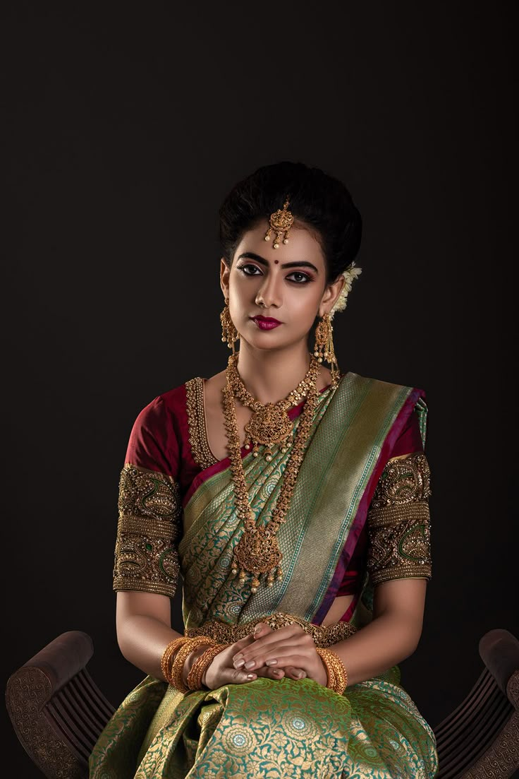 a woman in a green and gold sari sitting on a chair with her hands clasped