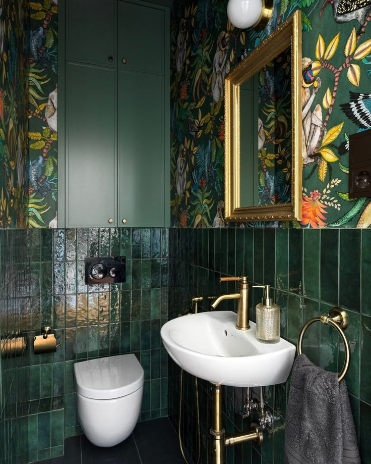 a green tiled bathroom with gold fixtures and floral wallpaper on the walls, along with a white toilet