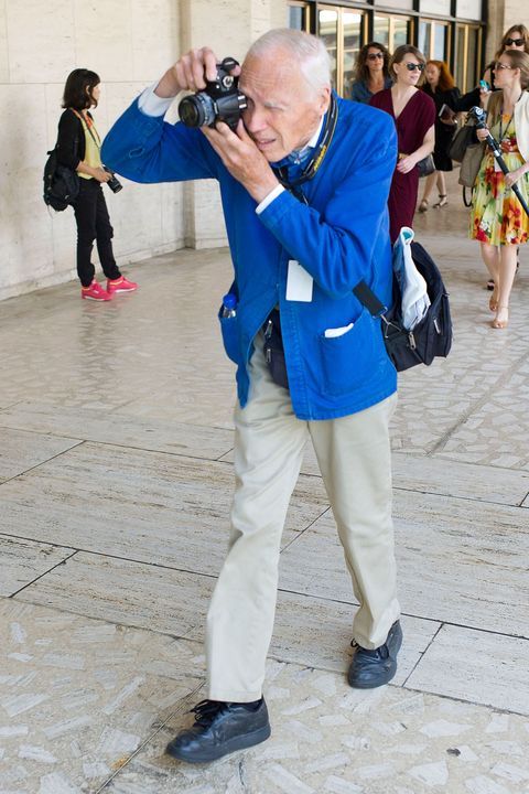 an older man taking a photo in front of a building with people walking around him