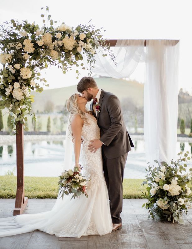 a bride and groom kissing under an arch decorated with flowers at the end of their wedding ceremony