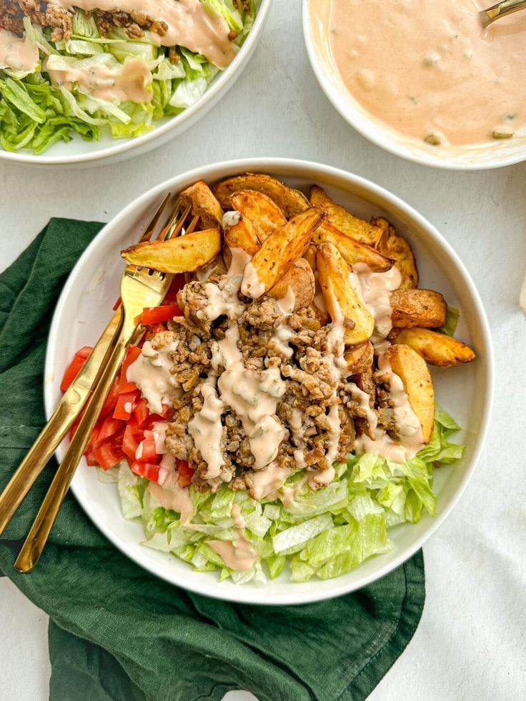 two white bowls filled with salad and fries on top of a green napkin next to some dipping sauce