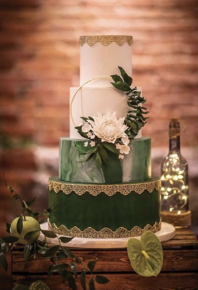 a three tiered green and white wedding cake with greenery on the top, next to a bottle of wine