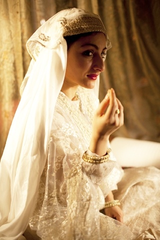 a woman in a white dress and veil sitting on a bed with her hands together