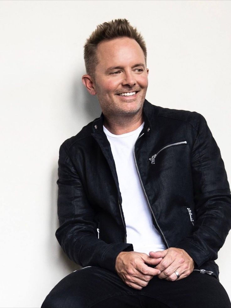 a man sitting on top of a white wall wearing a black jacket and smiling at the camera