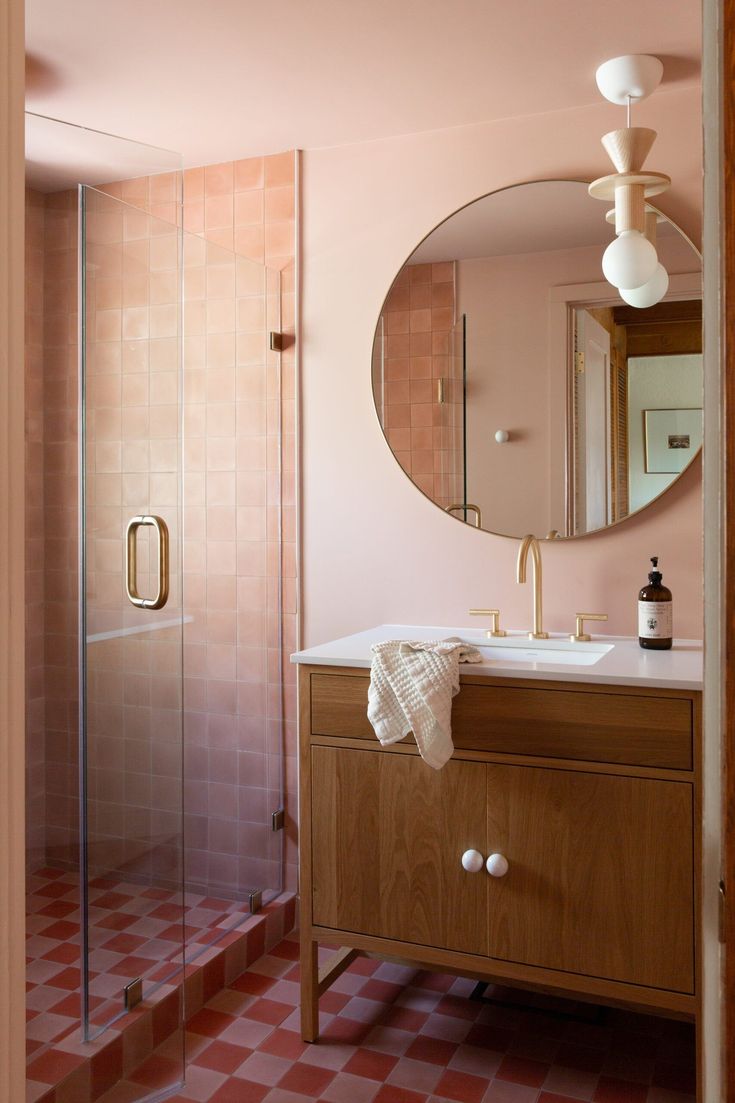 a bathroom with pink walls and checkered flooring, a round mirror above the sink