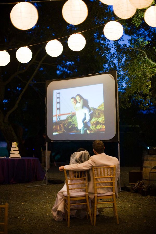 two people sitting at a table watching a movie on the screen with paper lanterns hanging above them