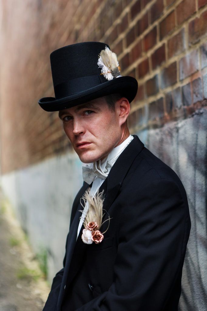 a man in a black suit and top hat leaning against a brick wall wearing a feathered boutonniere